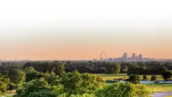 A panoramic view of a city skyline during sunset highlights the prominent arch and clustered high-rise buildings. In the foreground, lush green trees and open fields fill the landscape under a clear sky.