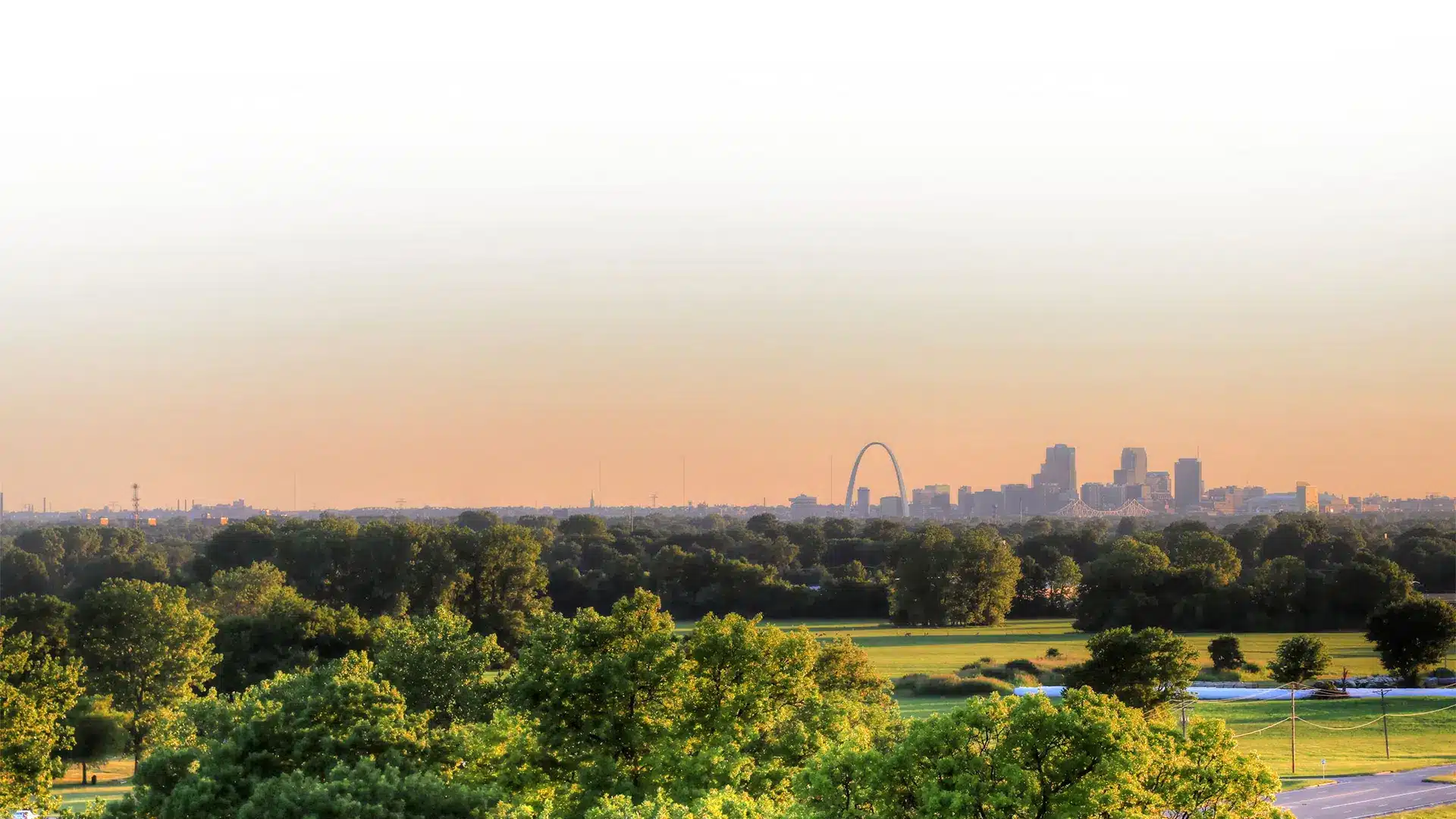 A panoramic view of a city skyline during sunset highlights the prominent arch and clustered high-rise buildings. In the foreground, lush green trees and open fields fill the landscape under a clear sky.