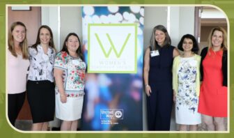 Six women stand together, smiling warmly in front of a sign for the Women's Leadership Society and United Way, a prominent non-profit organization. The backdrop displays a green "W" logo. They're dressed in business or semi-formal attire.