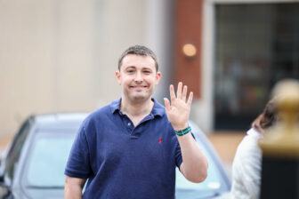 A person wearing a blue polo shirt is smiling and waving at the camera. He is outside with blurred buildings and a car in the background.